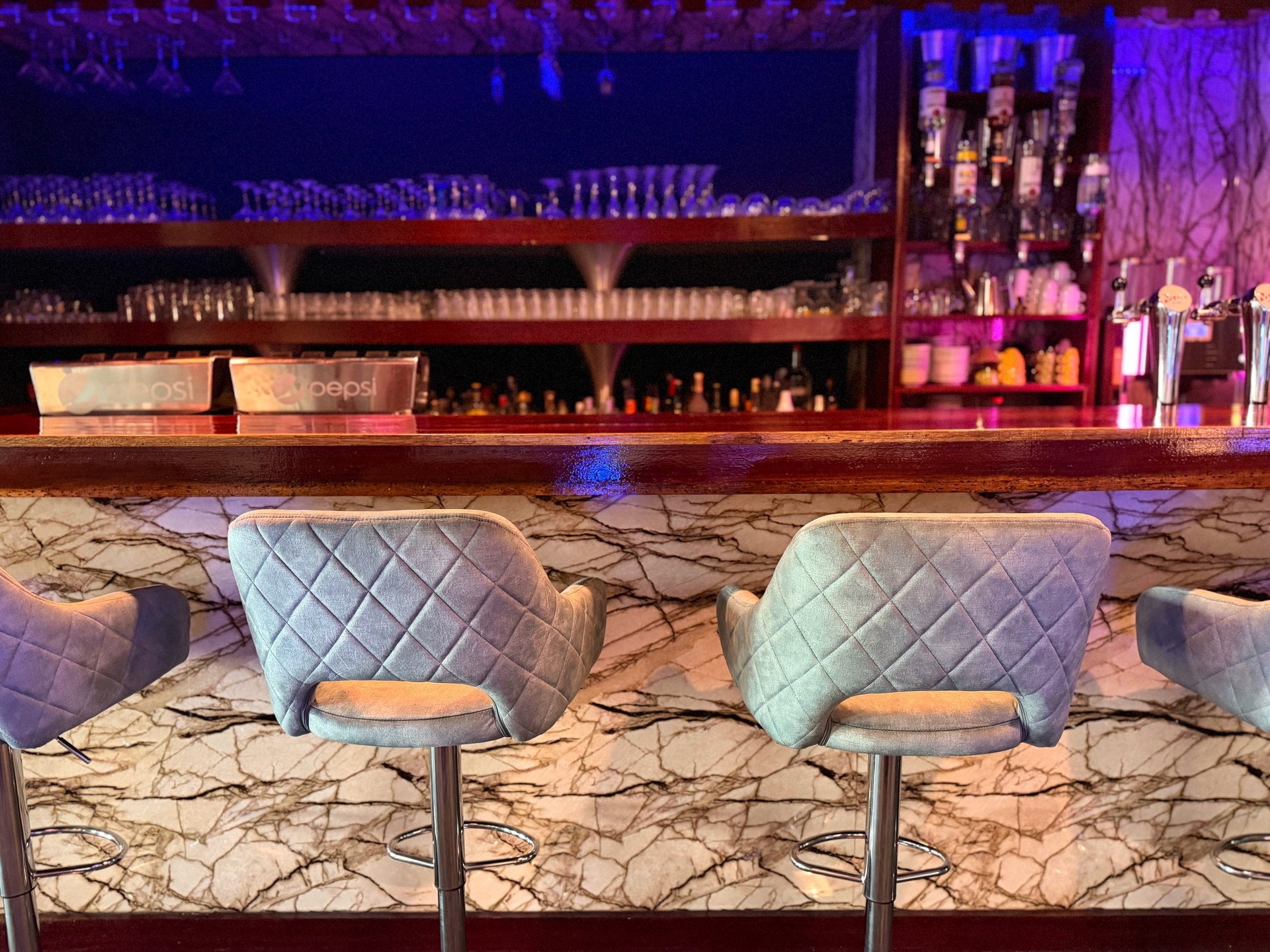 Modern bar seating with quilted gray chairs at a marble counter, illuminated bottles and purple ambient lighting